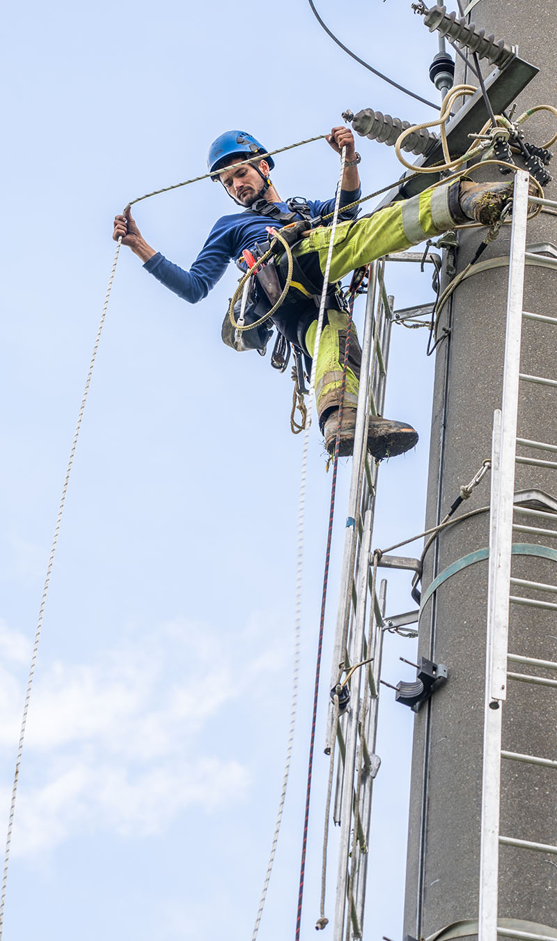 Netzelektiker hängt an Strommast und schaut in die Tiefe. 