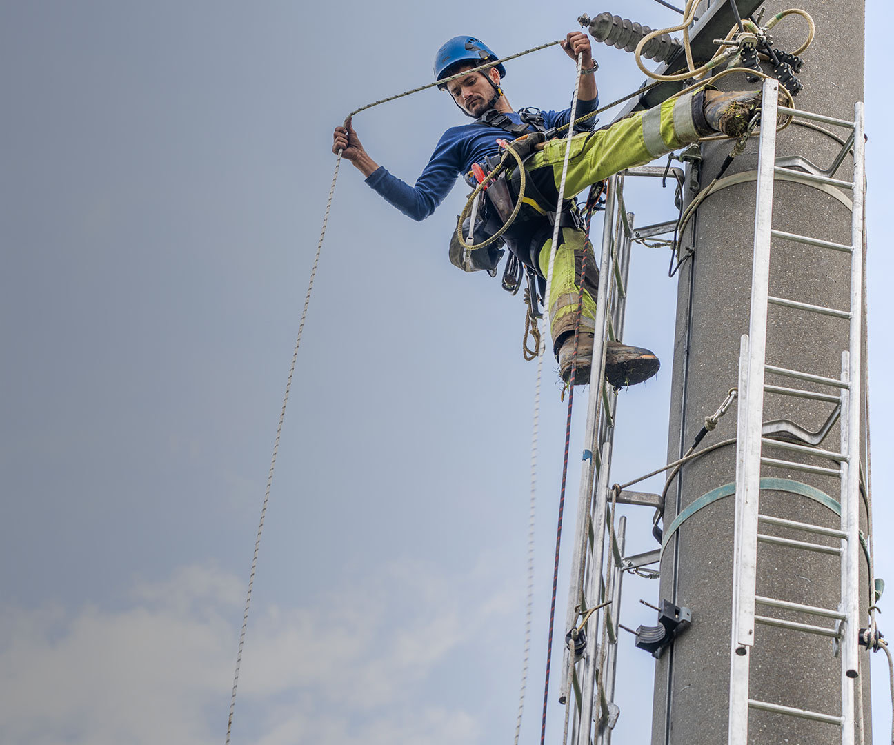 Netzelektiker hängt an Strommast und schaut in die Tiefe. 