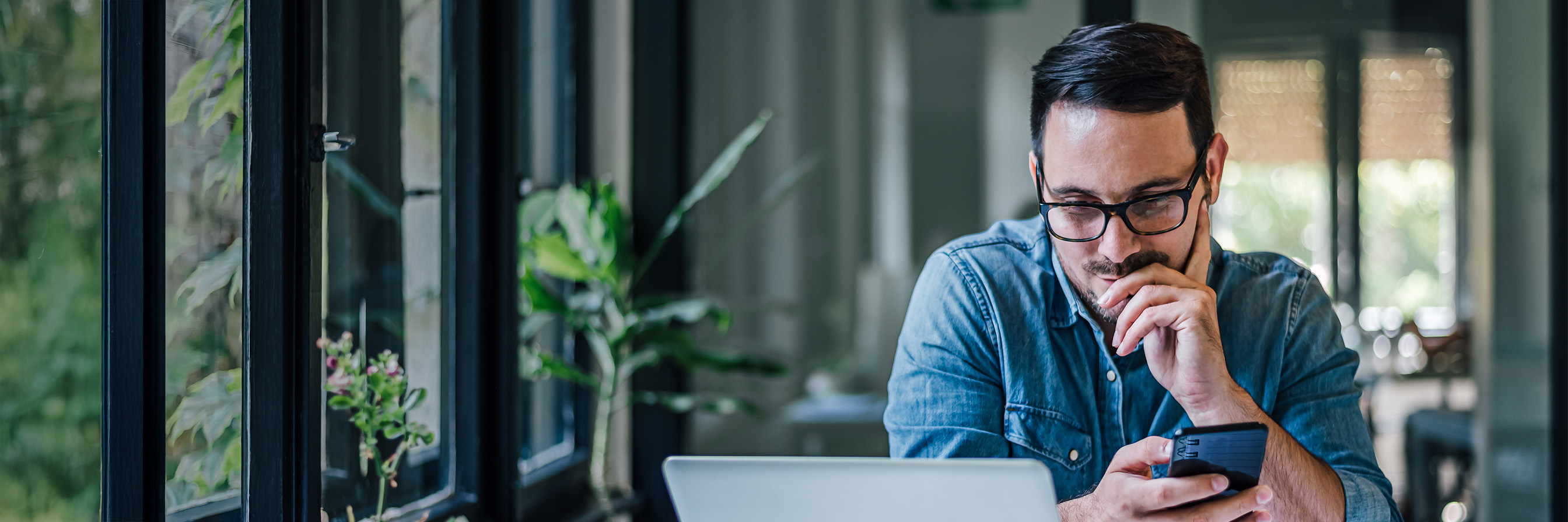 Mann an Laptop mit Mobiltelefon in der Hand. 