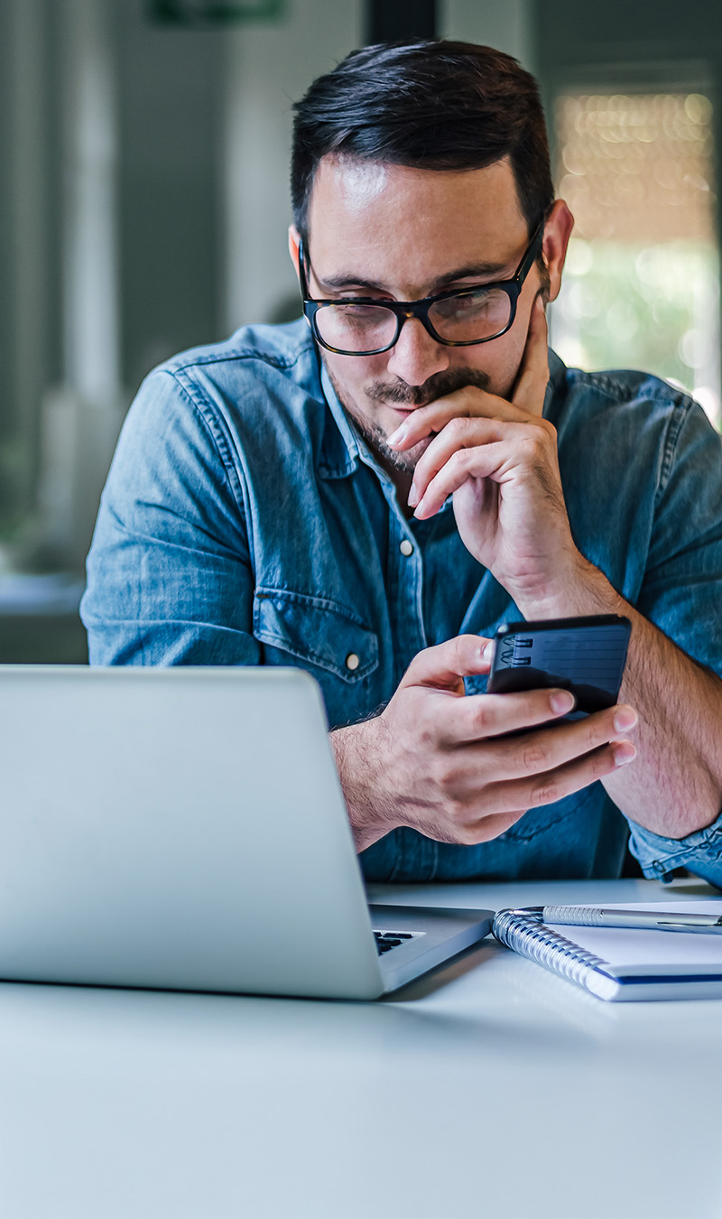 Mann an Laptop mit Mobiltelefon in der Hand. 