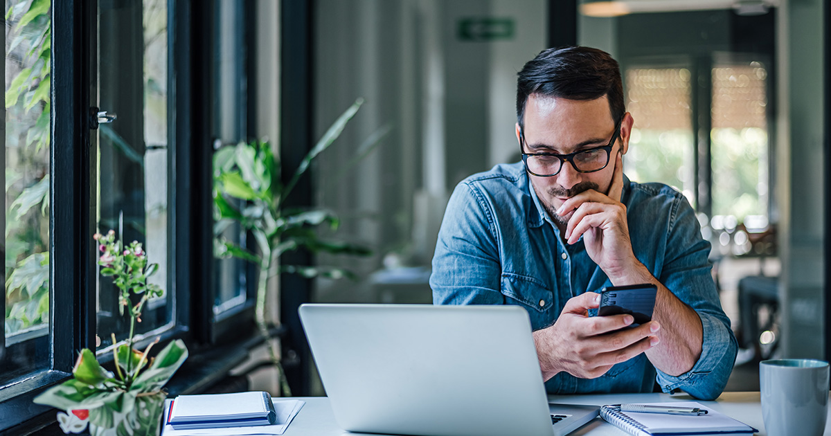 Mann an Laptop mit Mobiltelefon in der Hand. 