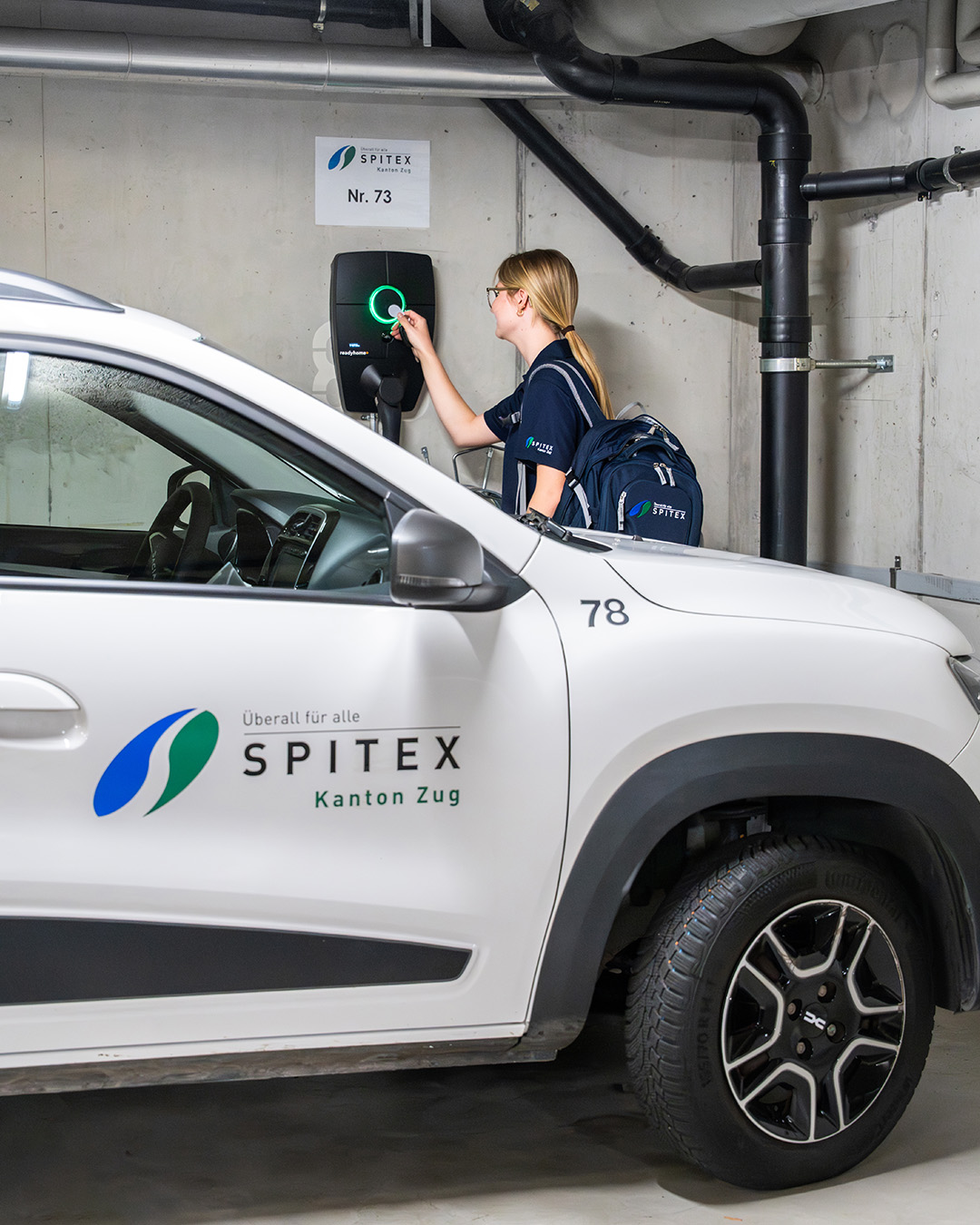 Pflegefachfrau steht hinter weissem E-Auto und bedient Ladestation. 